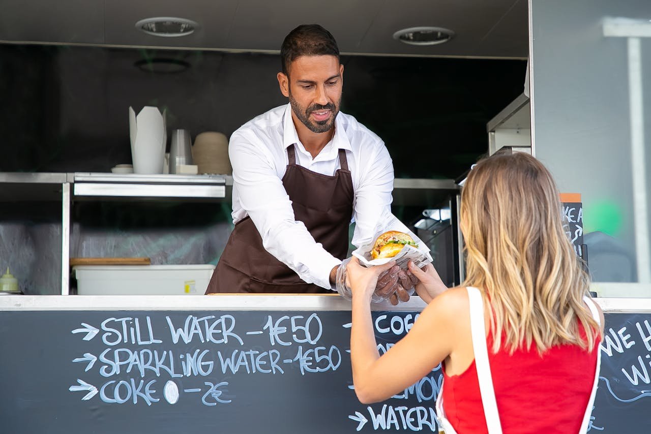home-hero Positive ethnic cook in apron standing at counter in food truck and giving delicious hamburger to anonymous woman customer in daytime